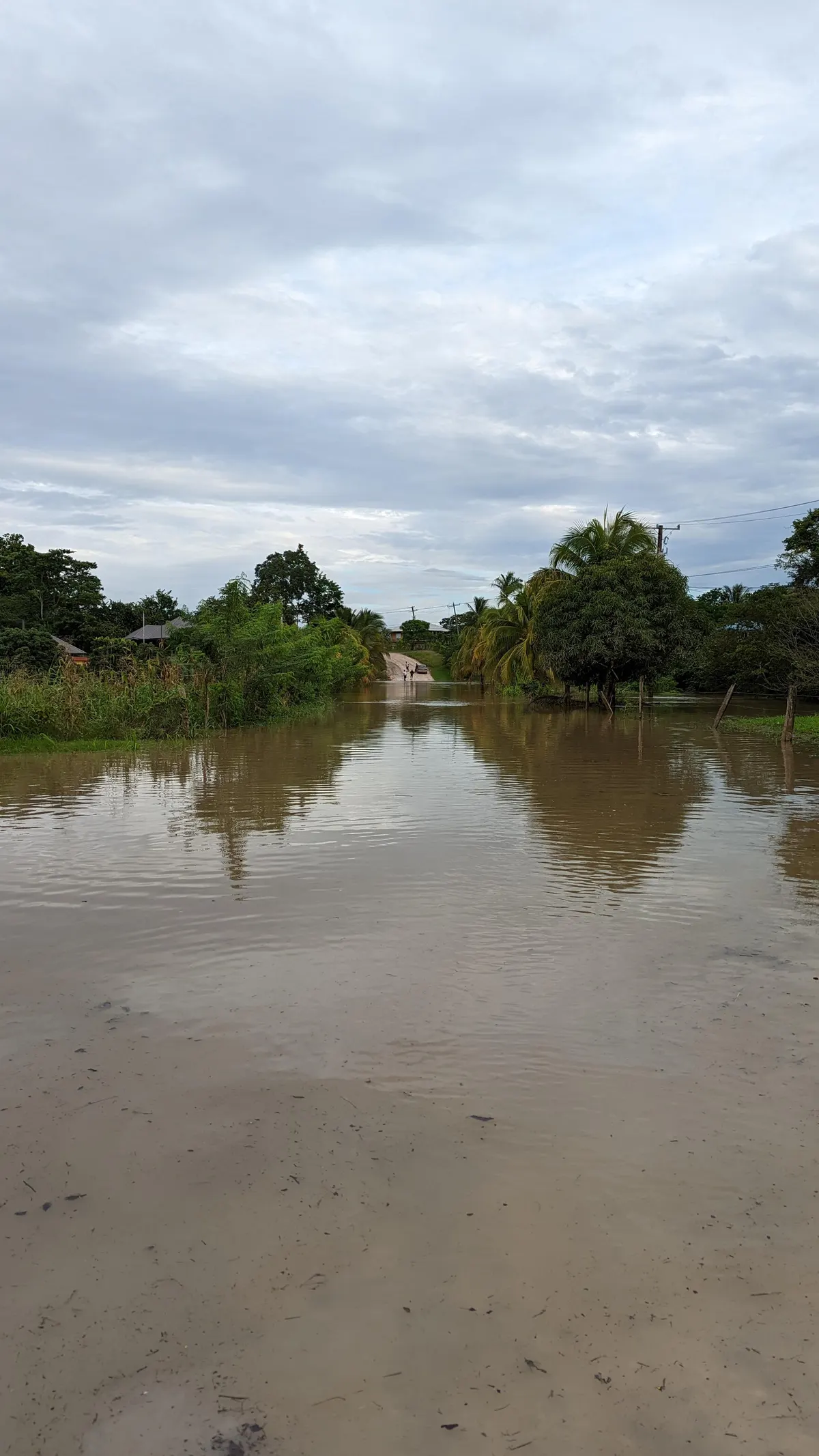 A flooded village road in Bullet Tree Falls, Cayo, Belize, with water covering the roadway and people wading in the distance