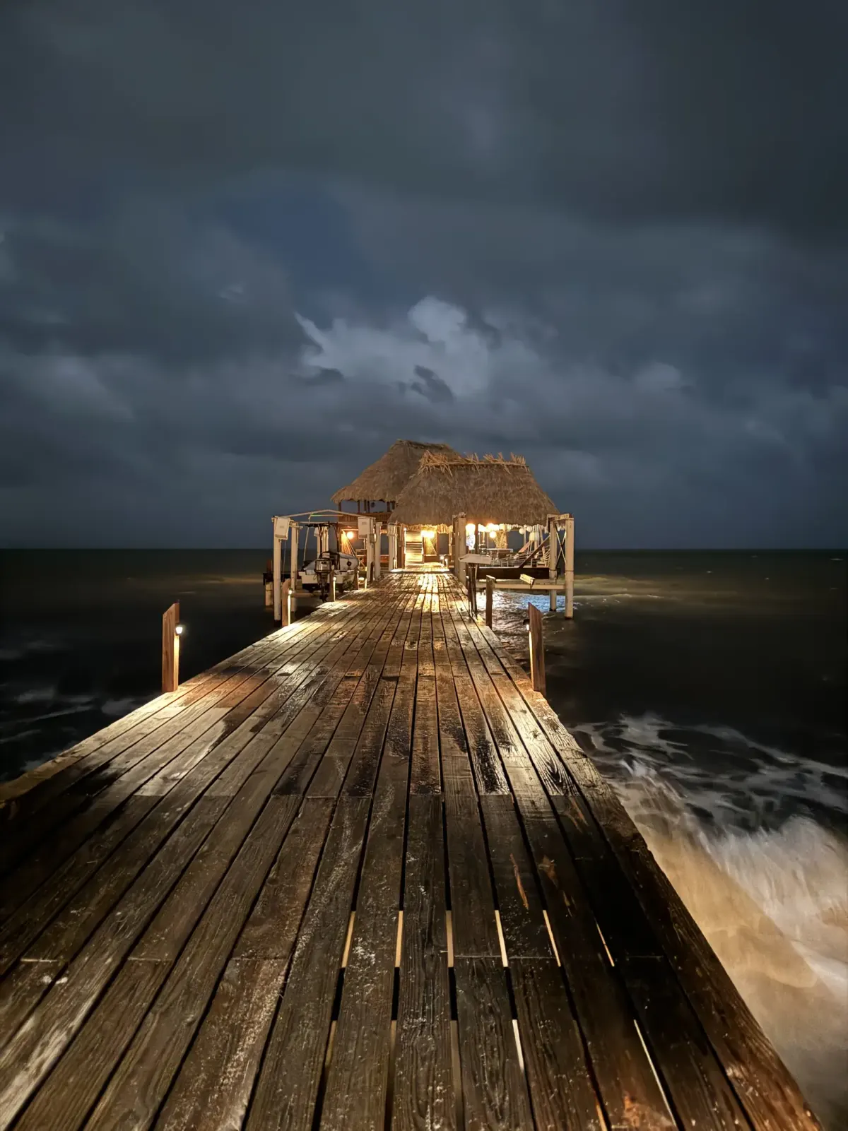 A wooden pier at dusk leading to a lit thatched palapa over the sea at Hopkins, Belize