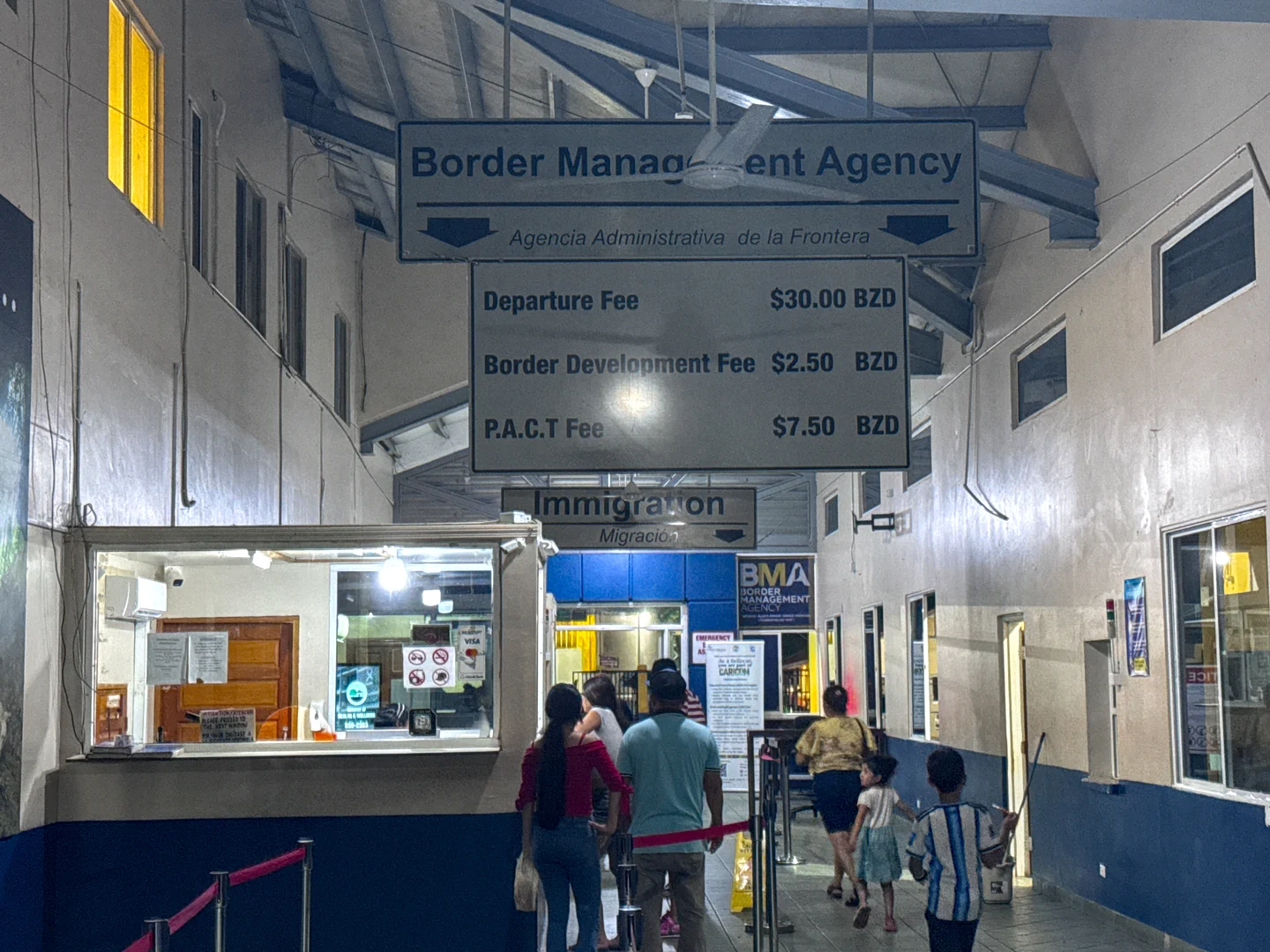 Border Management Agency fee board hanging above the immigration windows at the Belize border station, listing a BZ$30 departure fee, a BZ$2.50 border development fee, and a BZ$7.50 PACT fee