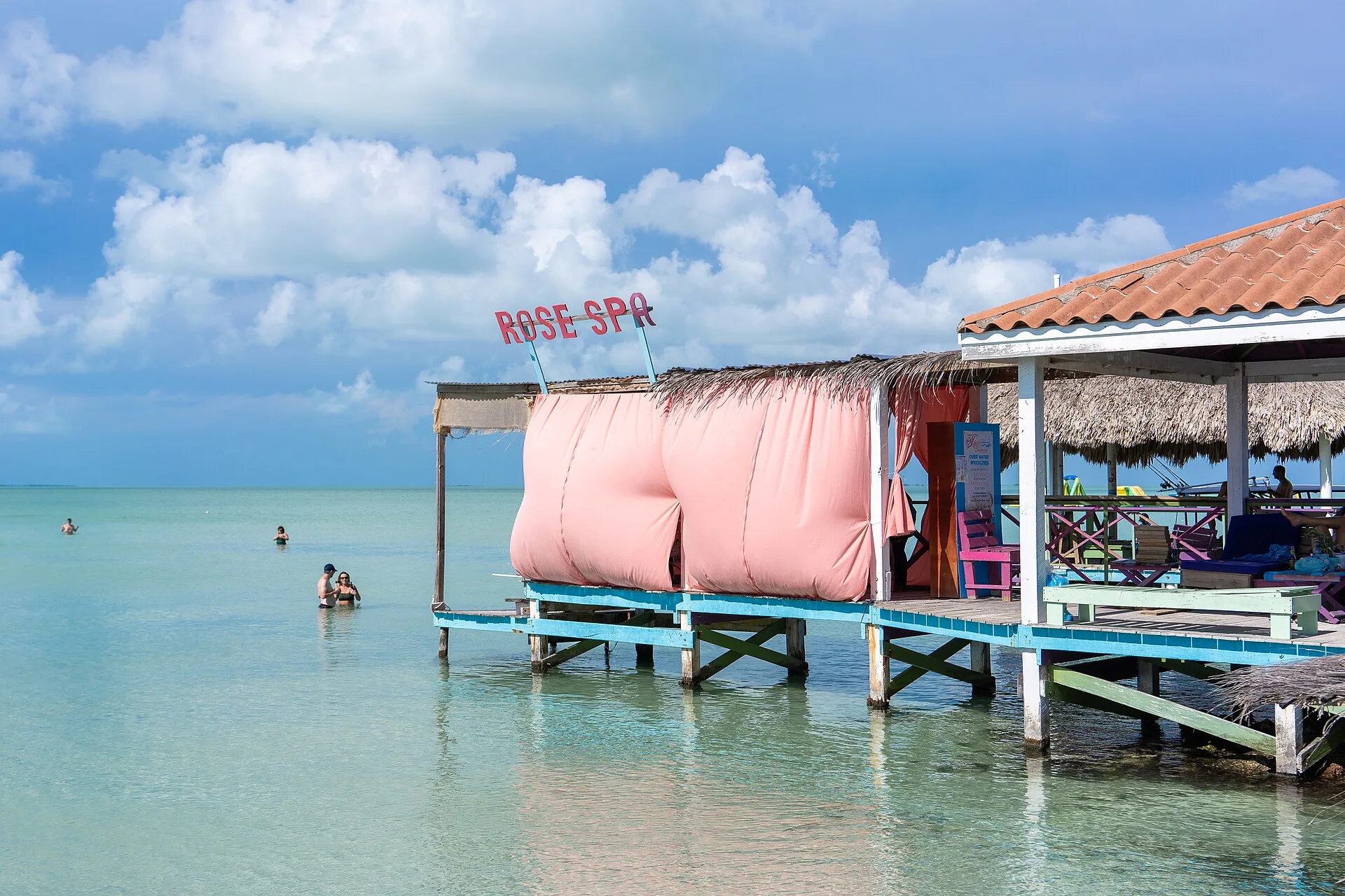 Swimmers standing in shallow turquoise water beside an over-water beach club at Secret Beach, Ambergris Caye