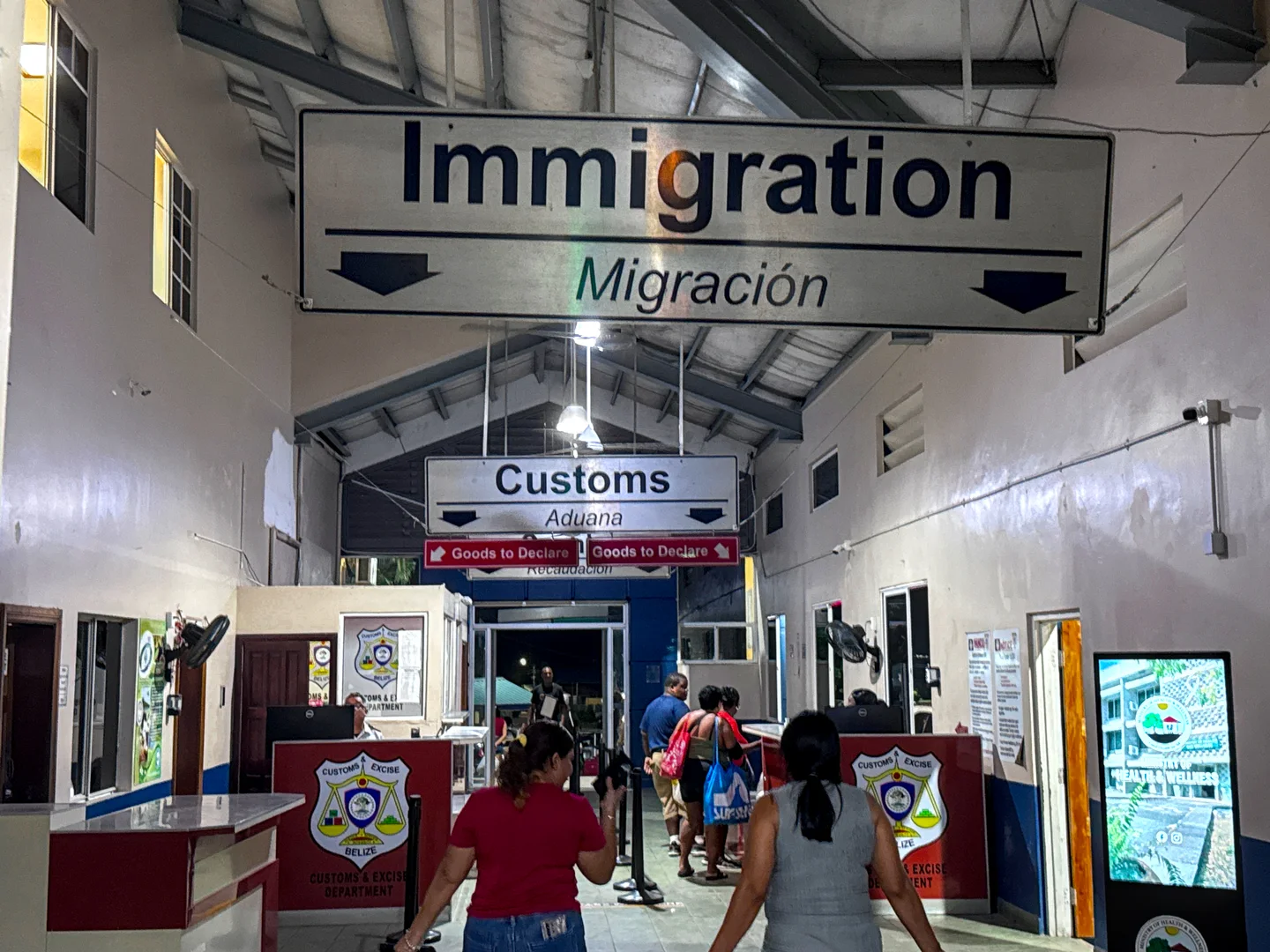 Interior of the Belize border station at Benque Viejo del Carmen, with overhead signs marking Immigration, Customs, Goods to Declare, and the cashier, and travelers queuing between rope barriers