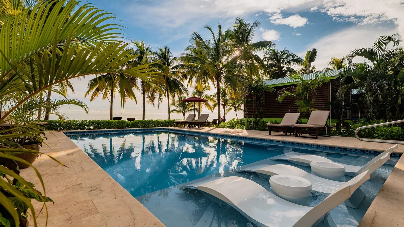 Poolside loungers at Coconut Row Boutique Hotel with a beachfront cabin and palm trees in the background at golden hour