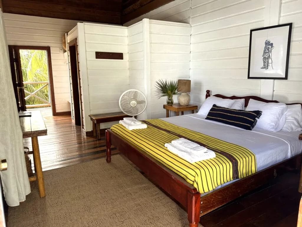 Guest room interior at Itza Resort with a queen bed dressed in a yellow striped throw, white plank walls, and a breezy porch doorway