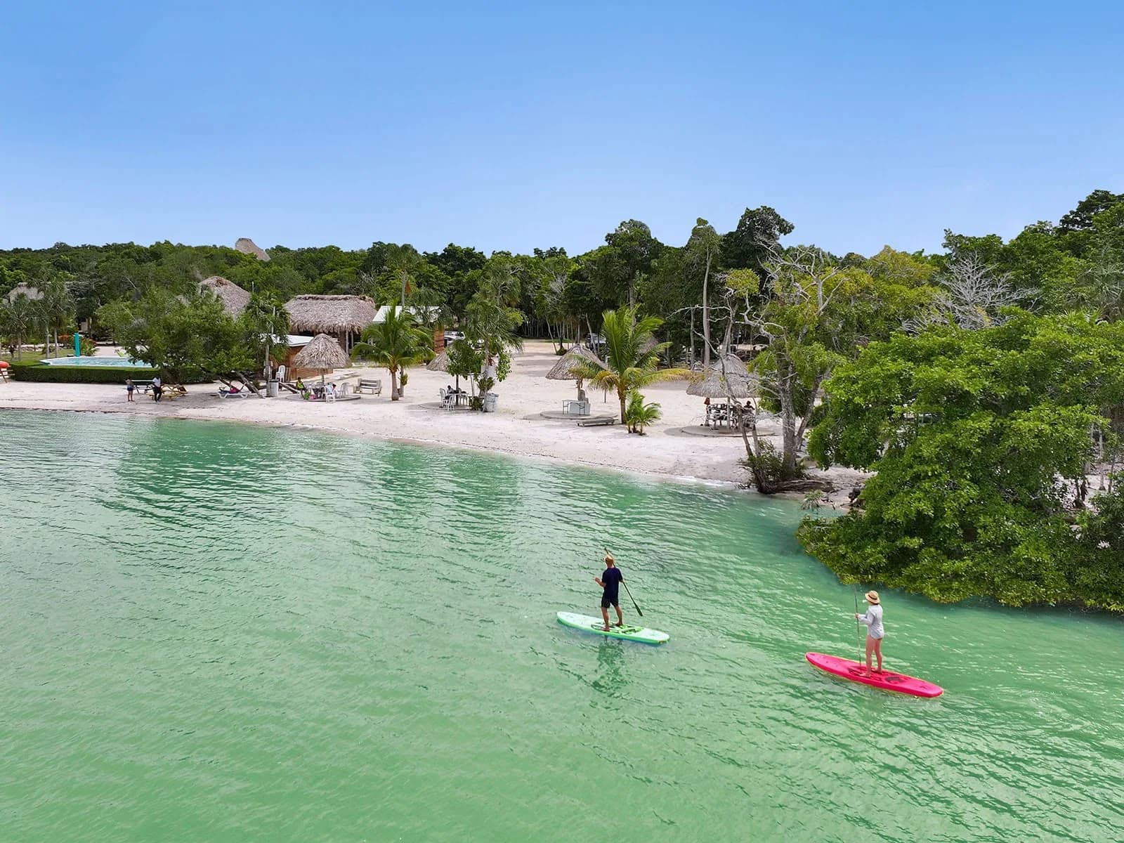 Guests paddleboarding off Orchid Bay Resort's beach, with the resort's pool and thatched palapas visible on shore