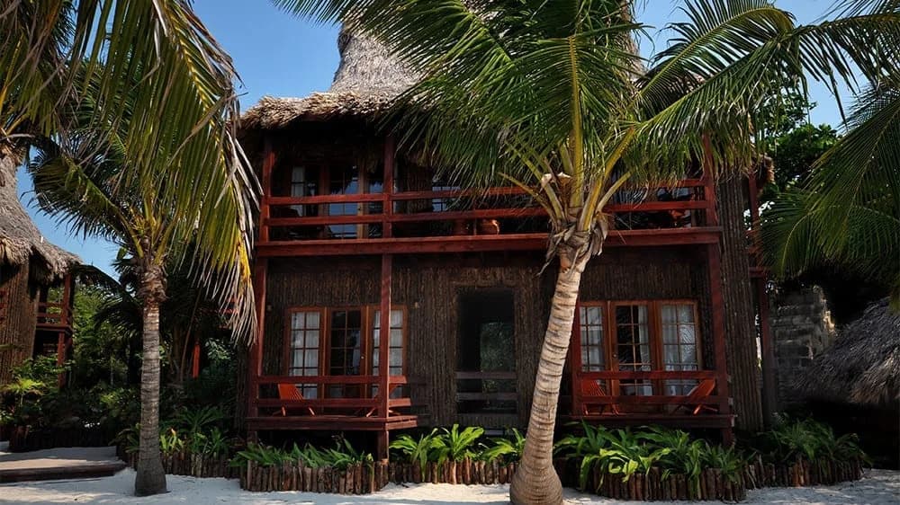 Exterior of a two-story thatched-roof cabana with private balconies and porches shaded by coconut palms at Ramon's Village Resort, Belize