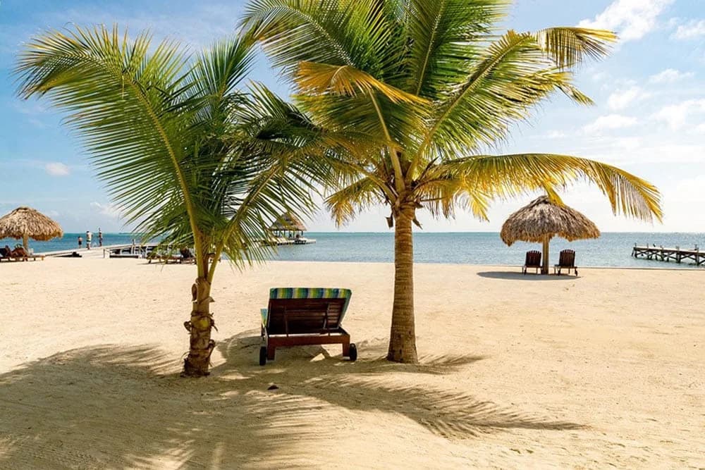 Palm shaded beach loungers and a thatched palapa on the sand at Xanadu Island Resort with the pier and Caribbean Sea in the background