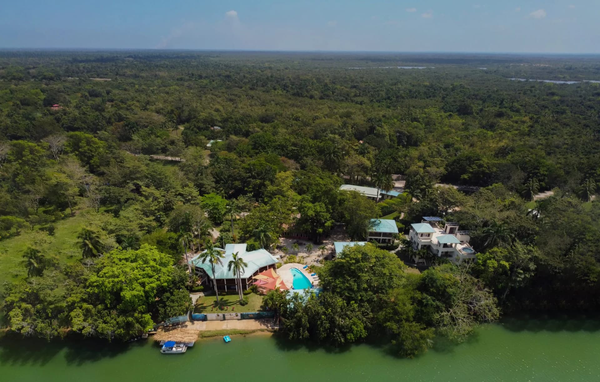 Aerial view of Black Orchid Resort's riverside jungle lodge complex on the Belize River, showing the main thatched-roof lodge, guest villas, a three-story building, and the pool deck.