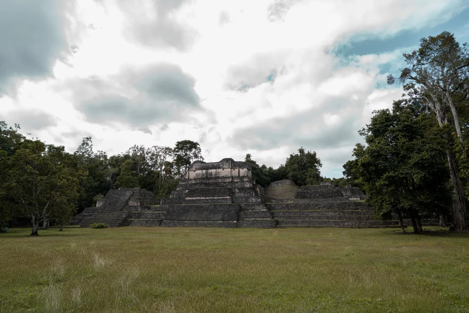Ancient Maya buildings at Caracol surrounded by rainforest beneath a dramatic cloudy sky.