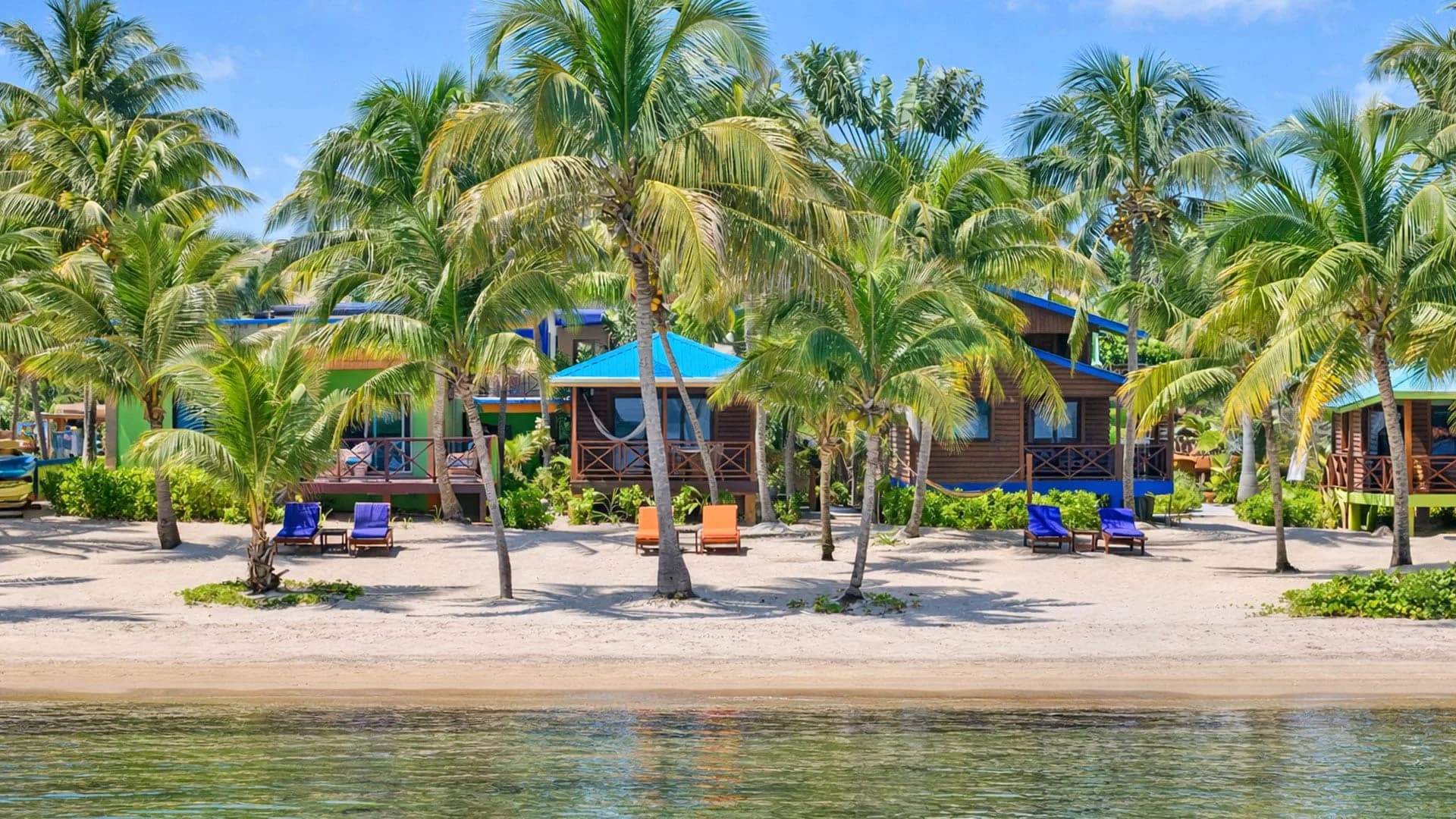 Row of Coconut Row Boutique Hotel's wooden beachfront cabins with green and blue roofs among palm trees on the sand in Hopkins, Belize