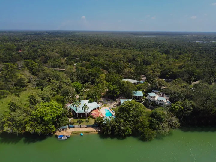 Aerial view of Black Orchid Resort's riverside jungle lodge complex on the Belize River, showing the main thatched-roof lodge, guest villas, a three-story building, and the pool deck.